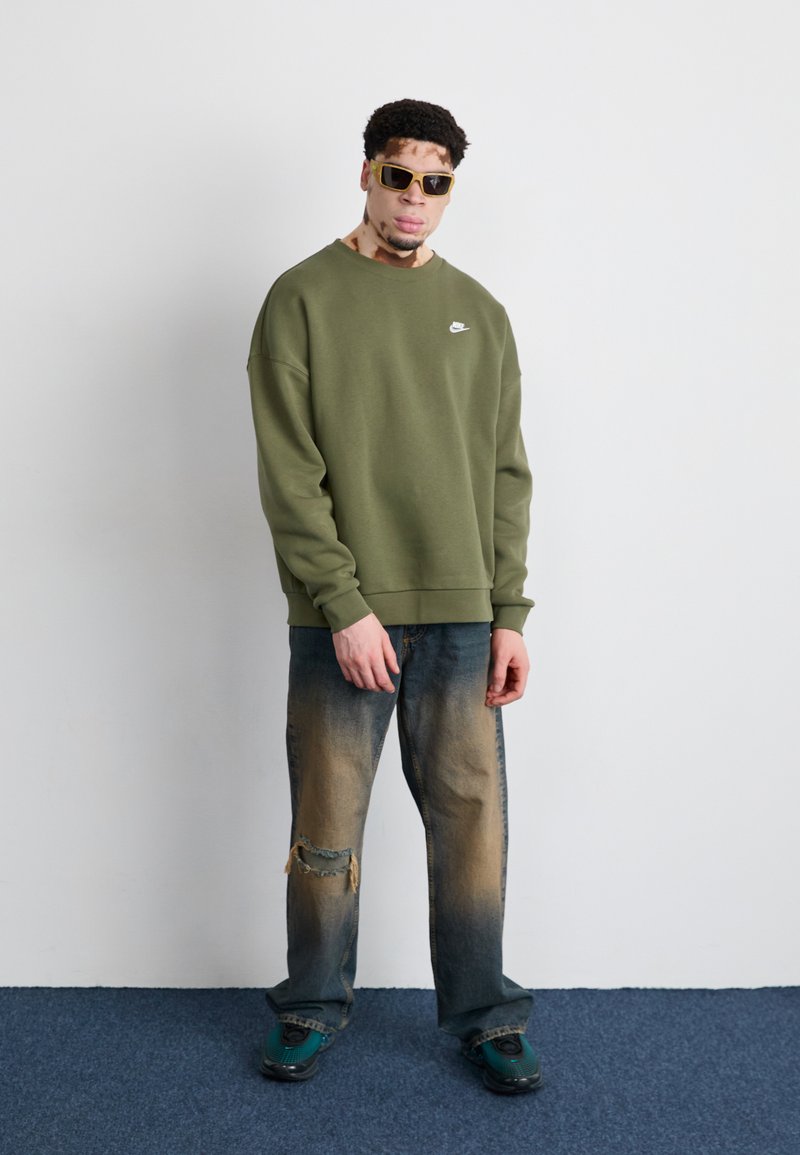 Felpa verde oliva con collo rotondo e logo bianco, abbinata a jeans blu strappati con orli sfrangiati. Scarpe da ginnastica verdi completano il look.