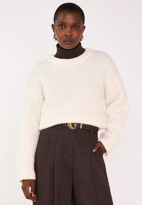 Maglione a coste color crema con scollatura ampia, indossato sopra un dolcevita marrone scuro, abbinato a pantaloni tailleur marroni a vita alta e una cintura.