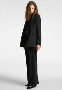Femme aux cheveux bouclés portant un blazer noir, un pantalon large et des chaussures pointues, regardant vers le bas devant un fond uni.