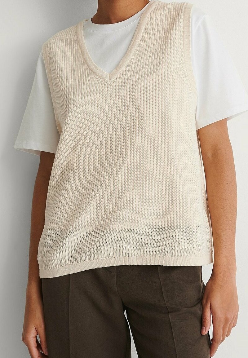 Personne portant un gilet sans manches en laine de couleur crème sur un t-shirt blanc à manches courtes et un pantalon marron foncé.