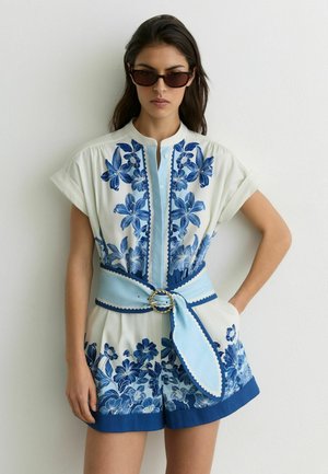 Kobieta w białym i niebieskim kombinezonie w kwiatowy wzór z szerokim pasem i ciemnymi okularami przeciwsłonecznymi, stojąca na tle jednolitej białej ściany.