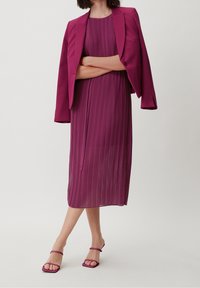 Mauve Plisseekleid, kombiniert mit einem passenden Blazer, der eine figurbetonte Taille und einen ausgestellten Rock hat. Vervollständigt mit mauvefarbenen Sandalen mit Riemchen.