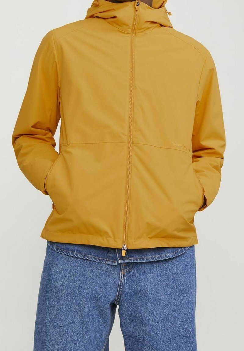 Homme portant une veste à capuche jaune moutarde avec fermeture éclair à l’avant, mains dans les poches, associée à un jean denim bleu.