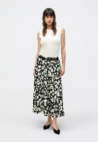 Gonna midi nera plissettata con motivo floreale bianco, abbinata a un top crema senza maniche e scarpe nere, con una cintura in vita.