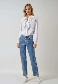 Blouse blanche à volants avec boutons, accompagnée d'un jean droit déchiré bleu clair et de baskets blanches. Tissu lisse et design décontracté.