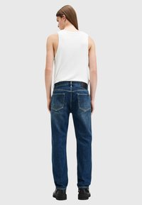 Los jeans de mezclilla azul tienen un corte recto, bolsillos traseros y un lavado claro, combinados con una blusa blanca sin mangas.
