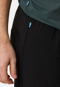 Svart sportshorts med en slät textur, med små svensknationalflagget-accenter på midjebandet och sidan, tillsammans med en grön mesh-tröja.