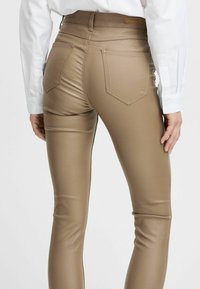 Pantalons en simili cuir métallisé beige, ajustés, avec poches arrière, texture lisse et taille mi-haute, associés à une chemise blanche.