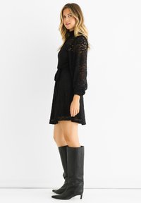 Black lace dress featuring long puffed sleeves, a fitted waist, and a flared skirt, paired with knee-high black leather boots.