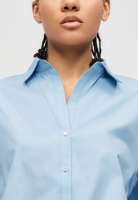 Personne aux cheveux tressés portant une chemise bleu clair boutonnée avec un col ouvert, sur un fond uni.