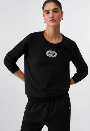 Mlada ženska, oblečena v črn dolg rokav s broško v obliki ovalnega dragulja na prsih in ujemajoče se črne hlače.