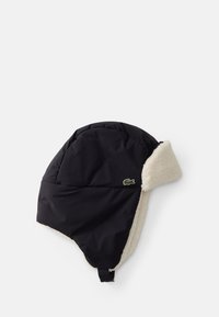 Cappello invernale imbottito di nero con fodera in pelliccia sintetica color crema, dotato di una patta laterale e un piccolo logo verde. Cinghia regolabile per una vestibilità sicura.