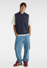 Gilet senza maniche blu navy con zip frontale, due tasche sul petto, indossato sopra una camicia a righe e abbinato a pantaloni cargo azzurri chiari e sneakers bordeaux.