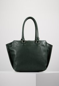 Ciemnozielona skórzana torebka na ramię z dwoma zaokrąglonymi uchwytami i delikatnym przeszyciem, stojąca na białej powierzchni na gładkim jasnym tle.