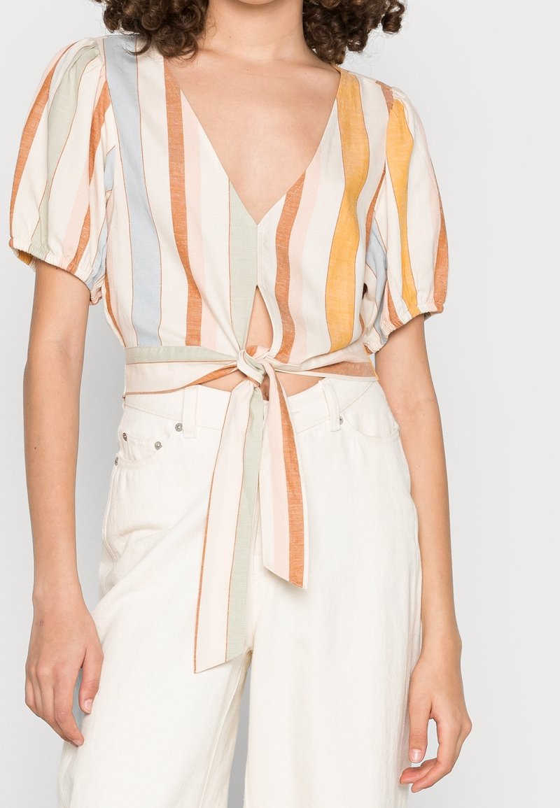 Femme portant une blouse rayée à manches courtes nouée à l'avant dans des tons pastel, associée à un pantalon blanc taille haute.