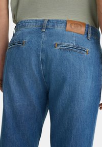 Jeans in denim blu con una finitura testurizzata, caratterizzati da un'etichetta in pelle marrone alla vita e due tasche posteriori con dettagli di cuciture arancioni.