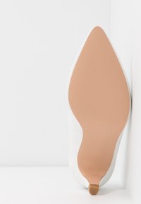 Chaussure blanche à talon haut avec un bout pointu et une tige lisse. Elle possède une semelle en caoutchouc brun clair à texture côtelée et un petit embout de talon.