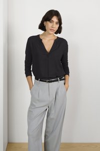 Cămașă neagră cu mâneci lungi, decolteu în V și detalii cu nasturi, asortată cu pantaloni gri deschis, cu talie înaltă și plii, și o curea neagră.
