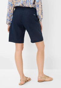 Shorts en lin bleu marine avec une taille élastique et une poche arrière, présentant une texture lisse et une coupe droite, associés à des sandales dorées.