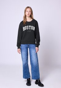 Czarna bluza z napisem "BOSTON 1980" w białym kolorze, w połączeniu z jasnoniebieskimi spodniami typu wide-leg i czarnymi, masywnymi butami. Gładka tekstura materiału.