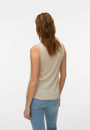 Kvinde med skulderlangt, bølget brunt hår iført en ærmeløs beige top og lyseblå jeans, stående vendt væk mod en ensfarvet baggrund.