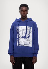 Μπλε φούτερ από μαλακό ύφασμα με γραφικό σχέδιο που απεικονίζει πουλί και αλυσίδα. Περιλαμβάνει κουκούλα με κορδόνι και ριμπ μανσέτες.