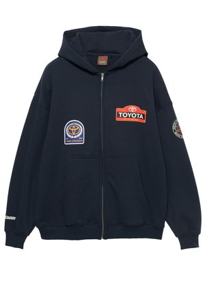 Tmavě modrá mikina s kapucí a celým zapínáním na zip, na přední straně a rukávu zdobená několika logy a nášivkami Toyota, s žebrovanými manžetami a lemem a kapucí se stahovací šňůrkou.