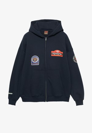 Tmavomodrá mikina s kapucňou na zips, zdobená viacerými logami Toyota a nášivkami na prednej strane a na rukáve, s rebrovanými manžetami a lemom a kapucňou so sťahovacou šnúrkou.