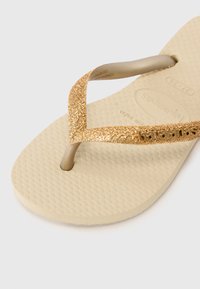 Beige-nahkasaappa, jossa on teksturoitu pohja ja kimaltavat kultaiset hihnat. Hihnoissa on logoyksityiskohta ja klassinen varvastossumalli.