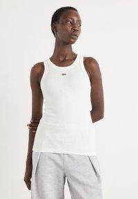 Hvit tanktop med ribbet tekstur og rund halslinning. Har en liten grønn Lacoste-logo på brystet. Bært med grå joggebukser.