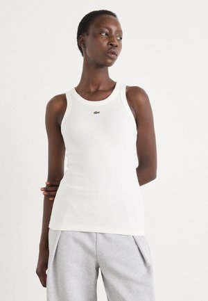 Débardeur blanc avec une texture côtelée et un col rond. Présente un petit logo vert Lacoste sur la poitrine. Porté avec un pantalon de jogging gris.