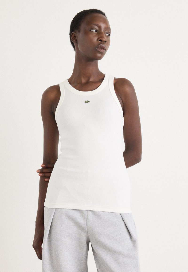 Hvit tanktop med ribbet tekstur og rund halslinning. Har en liten grønn Lacoste-logo på brystet. Bært med grå joggebukser.