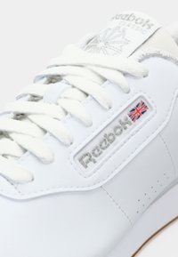 Sapatilha branca da Reebok com parte superior em couro texturizado, atacadores planos e etiqueta de logotipo com a bandeira do Reino Unido. Inclui detalhes de costura e perfurações.