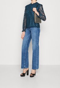 Petrolejová krajková halenka s dlouhými průsvitnými rukávy, kombinovaná s jasně modrými džíny s rovným střihem. Doplňky tvoří kovové psaníčko a černé platformové podpatky.
