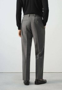 Persona vestida con pantalones grises a medida, mocasines negros y una blusa negra de manga larga, de pie frente a un fondo claro y liso.