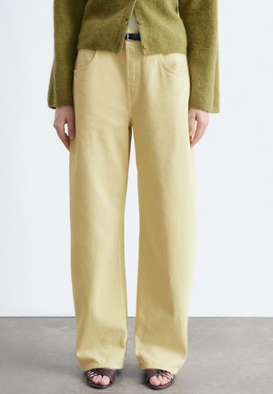 Person iført løstsiddende beige bukser, grøn knappet sweater, sort bælte og mørke åbne sandaler, stående på en grå gulv.