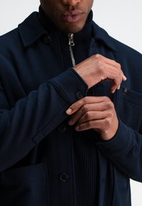 Marineblauwe jas met een knoopsluiting bij de kraag, voorzien van een stevige hoge nek met rits en een tekstuur gebreide onderlaag. Gladde stof met twee voorzakken.