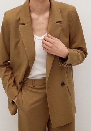 Person trägt einen beigefarbenen, doppelreihigen Blazer und passende Hose mit weißem, geripptem ärmellosem Top, eine Hand richtet den Blazer.