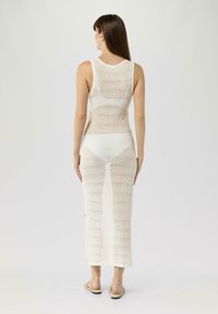 Robe en tricot sans manches, longueur genou, de couleur crème avec un motif en diamant. Présente une silhouette ajustée et des fentes sur les côtés. Portée avec des sandales à lanières.