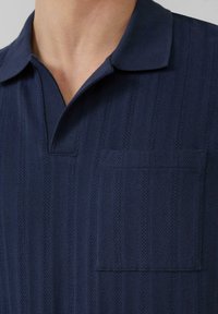 Navyblauwe polo met een ribbeltextuur en een visgraatpatroon. Beschikt over een zachte kraag en een klein borstzakje.