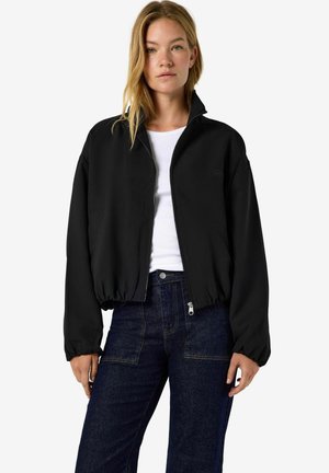 Femme portant une veste noire zippée par-dessus un haut blanc et un jean bleu foncé, debout devant un fond uni clair.