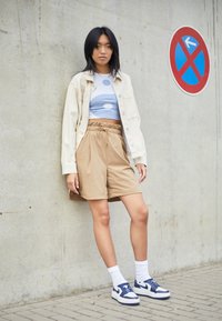 Ljusbeige oversized jacka över en blå crop top med cirkulära mönster, tillsammans med khaki shorts och vita strumpor med blå sneakers.