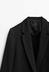 Musta bleiseri, jossa on teksturoitu kangas, viiltoetuja, yhdeksän nappia sulkeminen ja vuorattu sisäosa. Sisäpuolella näkyvä etiketti lukee "Massimo Dutti."