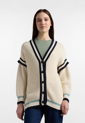 Nuori nainen pukeutuneena kermaiseen neulecardiganiin, jossa on mustia ja vaaleansinisiä raitoja, päällään vihreä toppi ja tummansiniset farkut valkoista taustaa vasten.