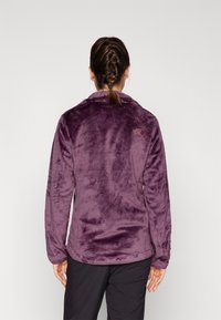 Kvinna med flätat hår som bär en lila fluffig jacka och svarta byxor, sedd bakifrån mot en enhetlig ljus bakgrund.