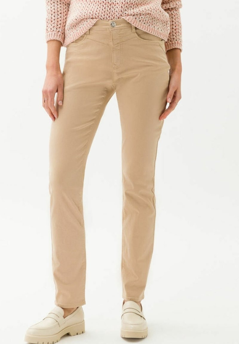 Pantalon ajusté couleur beige, confectionné dans un tissu lisse, doté d'une fermeture à boutons, de poches et d'une silhouette slim. Associé à des mocassins crème.