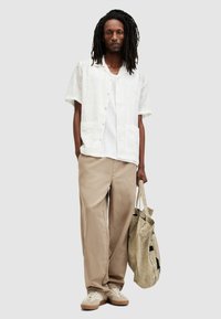 Camisa blanca de manga corta con botones, pantalones beiges holgados y zapatillas beiges, sosteniendo una bolsa de tela clara. Atuendo casual, con materiales texturizados.