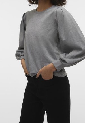 Kvinde iført en grå langærmet bluse med puffede skuldre og sorte bukser, med hænderne gemt i forlommerne.