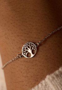 Zilveren armband met een ronde hanger met een ingewikkeld boomontwerp, rustend op de huid met zichtbare textuur. Kettinglinkdetails zijn zichtbaar.