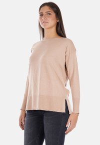 Maglione a maglia beige chiaro con maniche lunghe, scollatura tonda, spacchi laterali all'orlo e una vestibilità rilassata. Indossato con jeans scuri.